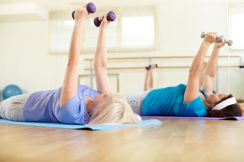 Zwei Frauen liegen auf Gymnastikmatten und heben Gewichte in einer Fitnessumgebung.