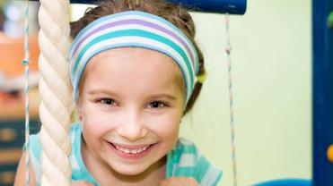 Lächelndes Mädchen mit blauer Kopfband und gestreifter Bluse hält sich an einem Klettergerüst fest.