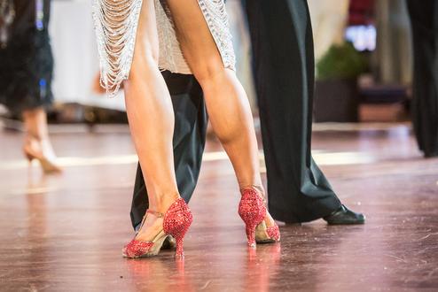 Nahaufnahme von tanzenden Füßen, eine Person trägt glitzernde rote Tanzschuhe auf einem schimmernden Parkettboden.