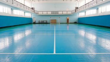 Leerer Sporthalle mit blauer Bodenfläche, seitlichen Wänden und Tor auf der gegenüberliegenden Seite.